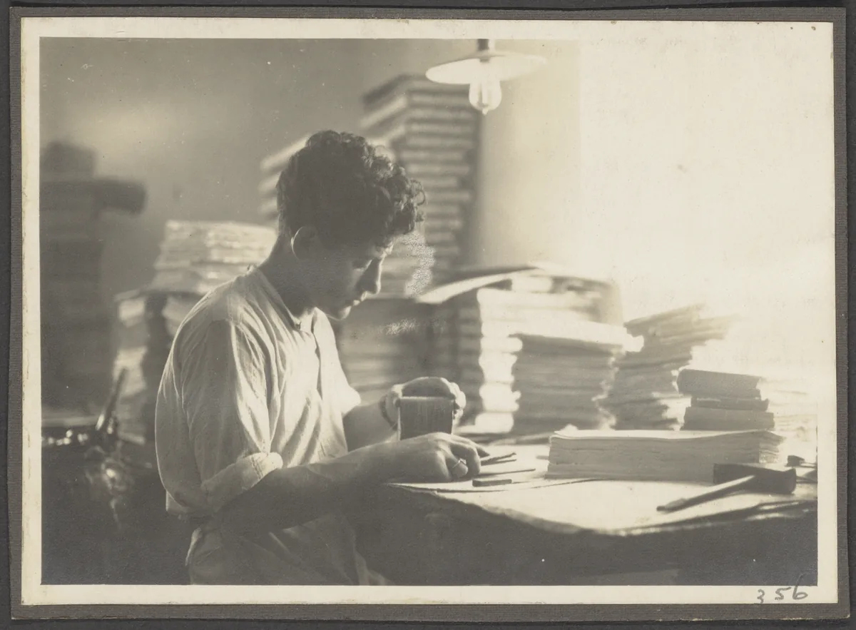 Sepia photograph of a young man working at a desk surrounded by tall stacks of paper, under a hanging lamp.