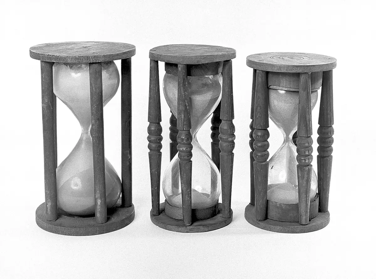 Three antique hourglasses in turned wooden frames, of slightly different sizes, photographed in black and white.
