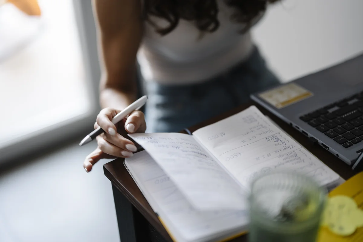 A person sitting at a desk writing notes by hand in a lined notebook, an open laptop beside them.