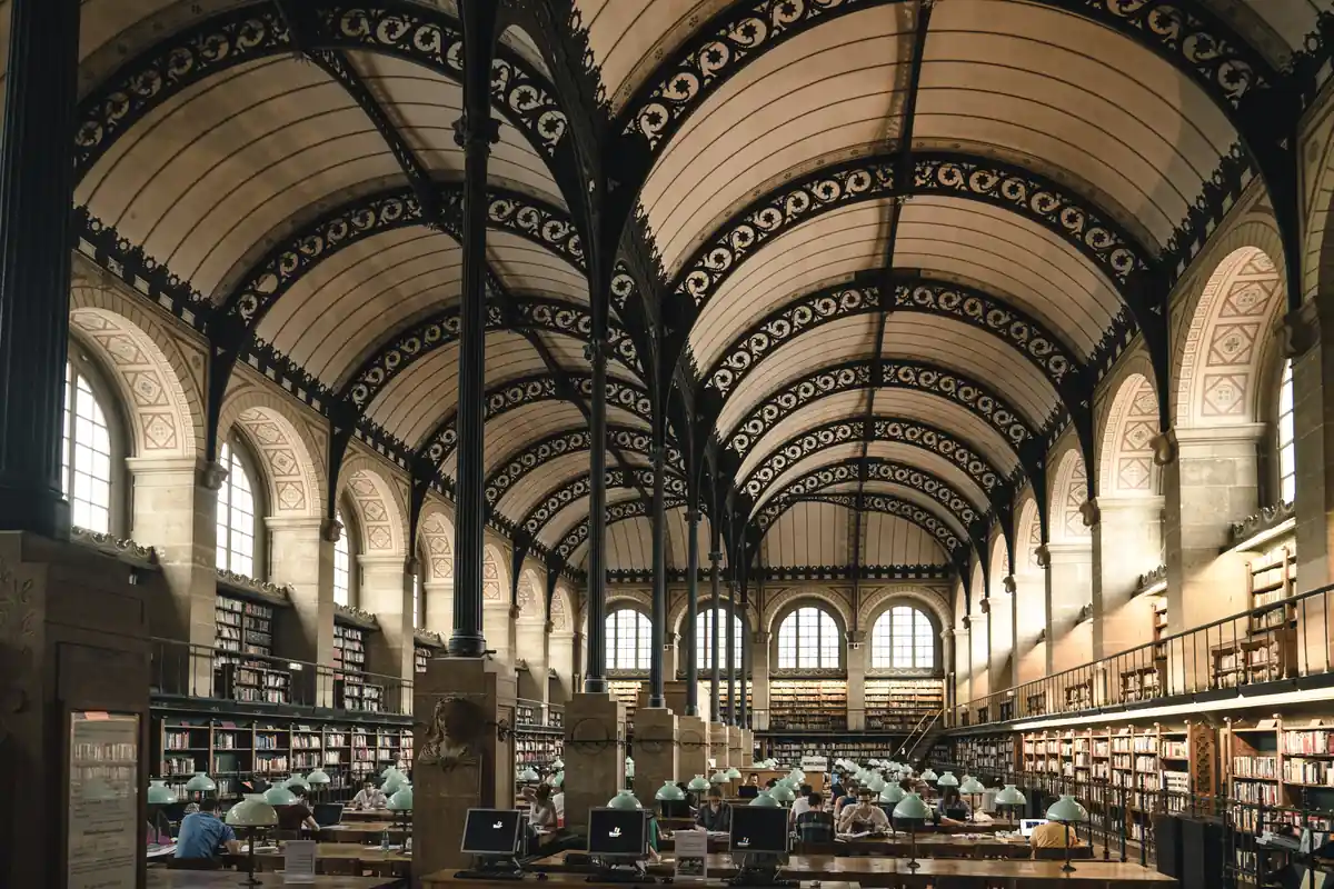 The vaulted reading room of the Bibliotheque Sainte-Genevieve in Paris, rows of desks with green lamps beneath iron arches, lined with bookshelves.