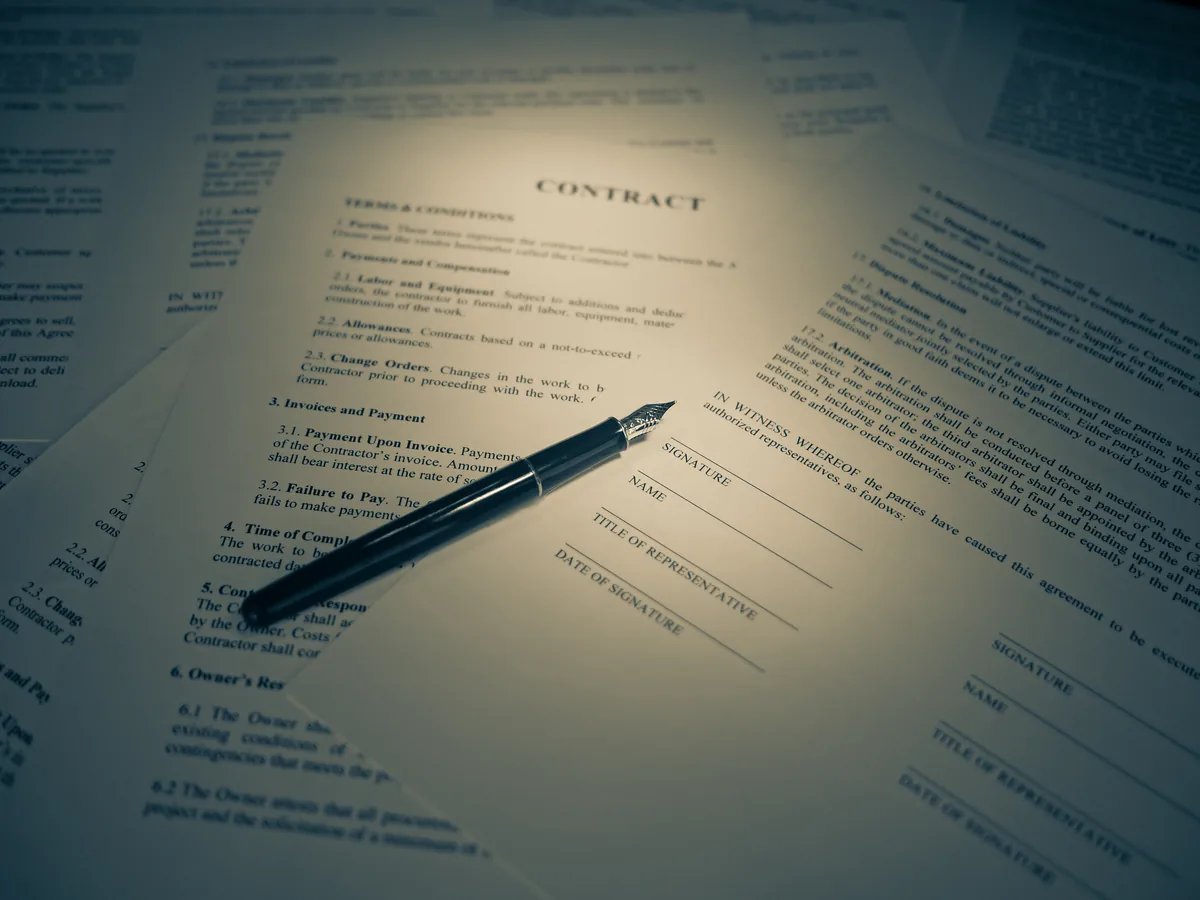 Printed contract pages spread across a desk under a pool of lamplight, with a fountain pen resting on the signature block.